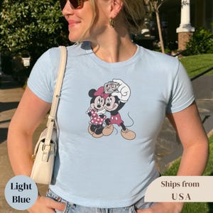 Pu&ograve; includere: T-shirt azzurra con una grafica di Topolino e Minni che si fanno una foto. Minnie indossa un vestito e un fiocco a pois rosa. Topolino indossa pantaloncini rossi. La maglietta ha le parole "Light Blue" in basso a sinistra.