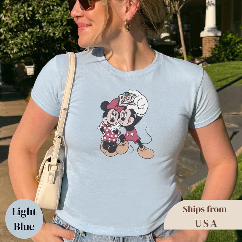Pu&ograve; includere: T-shirt azzurra con una grafica di Topolino e Minni che si fanno una foto. Minnie indossa un vestito e un fiocco a pois rosa. Topolino indossa pantaloncini rossi. La maglietta ha le parole "Light Blue" in basso a sinistra.
