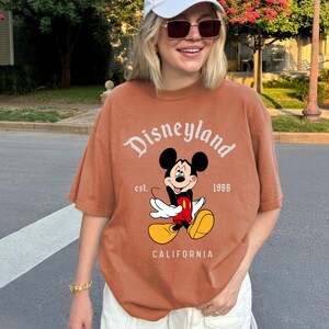 May include: A rust-colored t-shirt featuring the Disneyland logo and a graphic of Mickey Mouse. The text "est. 1955" and "CALIFORNIA" are also printed on the shirt. The shirt has short sleeves and a relaxed fit.