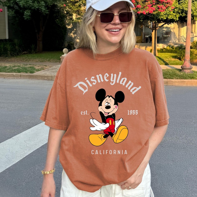 May include: A rust-colored t-shirt featuring the Disneyland logo and a graphic of Mickey Mouse. The text "est. 1955" and "CALIFORNIA" are also printed on the shirt. The shirt has short sleeves and a relaxed fit.