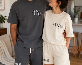 May include: A charcoal grey t-shirt with the text Mr. and matching shorts with the text Smith, alongside a cream t-shirt with the text Mrs. and matching shorts with the text Smith. Both sets feature a simple crew neck and a relaxed fit design.