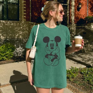 May include: A green t-shirt featuring a black graphic of Mickey Mouse. The shirt is a solid color and has a relaxed fit. The image shows a person wearing the shirt.