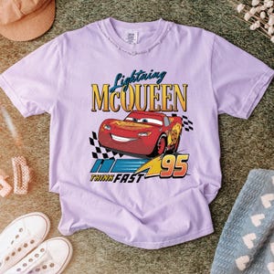 May include: A light purple t-shirt featuring Lightning McQueen from the Cars movie. The graphic includes the text "Lightning McQueen", the number "95", and the phrase "Think Fast". The design is in red, yellow, and blue.
