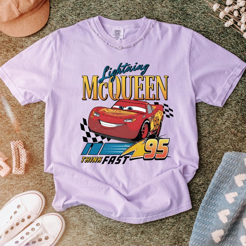 May include: A light purple t-shirt featuring Lightning McQueen from the Cars movie. The graphic includes the text "Lightning McQueen", the number "95", and the phrase "Think Fast". The design is in red, yellow, and blue.