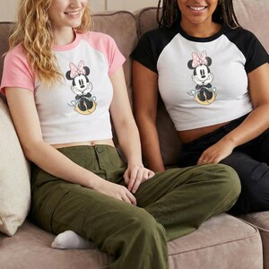 May include: White crop top with a Minnie Mouse graphic, featuring a pink bow and yellow accents. The shirt has pink raglan sleeves. The model is wearing olive green cargo pants.