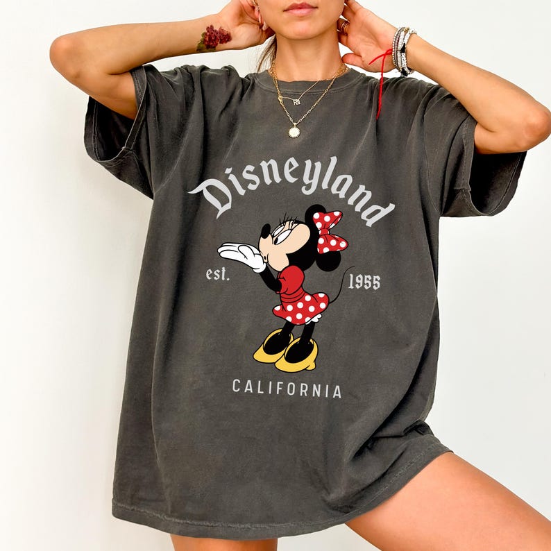 May include: Dark gray t-shirt featuring Minnie Mouse in a red polka dot dress and bow. The shirt has the Disneyland logo, the text "est. 1955", and "CALIFORNIA" printed on it. The shirt is a relaxed fit.