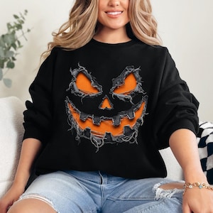 May include: A black long-sleeve sweatshirt featuring a large graphic of a jack-o'-lantern face. The design displays orange eyes, nose, and a wide, jagged mouth with a distressed, frayed fabric effect layered over the black material.