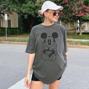 May include: Dark gray t-shirt featuring a black graphic of Mickey Mouse with his hands under his chin. The shirt has short sleeves and a relaxed fit. The person in the image is wearing a white cap and sunglasses.