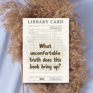 May include: A beige library card with the text "What uncomfortable truth does this book bring up?" is displayed against a backdrop of dried ornamental grass and a light blue surface. The card has a vintage aesthetic.