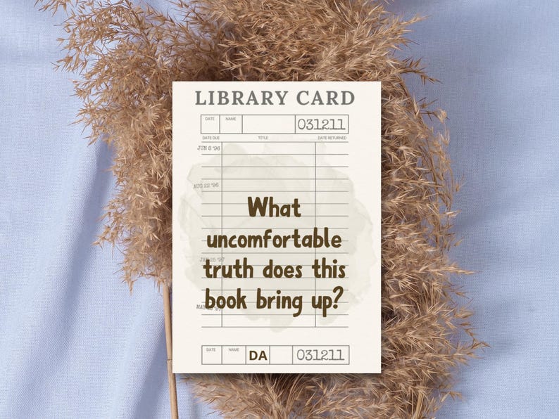 May include: A beige library card with the text "What uncomfortable truth does this book bring up?" is displayed against a backdrop of dried ornamental grass and a light blue surface. The card has a vintage aesthetic.