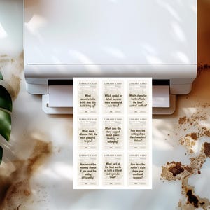 May include: A white printer printing nine library cards with text. The cards have a vintage aesthetic with questions like "What uncomfortable truth does this book bring up?" and "Which symbol or detail becomes more meaningful over time?"