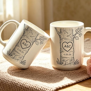 Peut inclure: Deux tasses en céramique blanche ornées d'un dessin au trait noir représentant un tronc d'arbre avec un cœur gravé dans l'écorce. À l'intérieur du cœur figurent les lettres A&C et la date 6.26.26. Une tasse est droite, l'autre est inclinée.