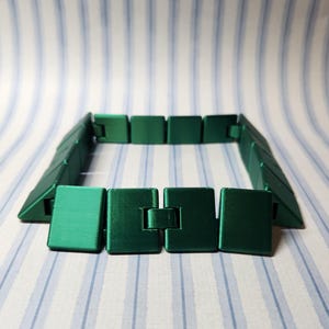 May include: A green bracelet with a geometric design. The bracelet is made of square and triangular links that connect to form a flexible band. The clasp is a simple, rectangular design.