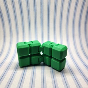 May include: Two green infinity cubes are displayed against a backdrop of white and blue striped fabric. The cubes are connected and appear to be made of plastic. The cubes are a bright, solid green color.