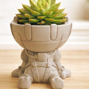 May include: A light grey astronaut-shaped planter holding a green succulent plant. The planter has detailed features of an astronaut suit. The succulent has green leaves with reddish tips. The planter is sitting on a wooden surface.