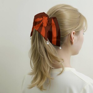Op de afbeelding: Een haaraccessoire met een strik in afwisselend donkerbruine en feloranje verticale strepen. De strik is bevestigd aan een haarelastiek en is versierd met drie kleine, ronde, witte kunstparels die aan de onderranden van de stof hangen.