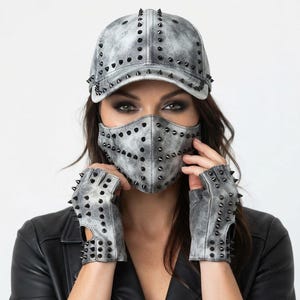Puede incluir: Una gorra de b&eacute;isbol, una mascarilla y guantes sin dedos con un acabado gris y negro desgastado. Cada art&iacute;culo presenta una textura envejecida y est&aacute; adornado con numerosos remaches met&aacute;licos negros y afilados dispuestos en un patr&oacute;n de rejilla.