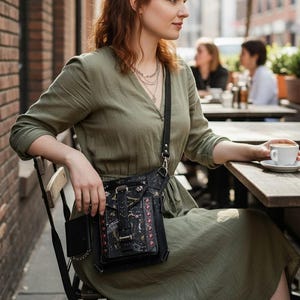 May include: A black crossbody bag with a decorative design and a small side pocket. The bag has a buckle closure and a long strap. The person is wearing a green dress and sitting at a wooden table with a cup of coffee.