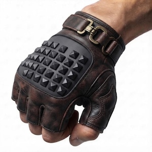 May include: A pair of fingerless gloves crafted from dark brown distressed leather, featuring a prominent black rectangular panel on the back of the hand adorned with a grid of pyramid-shaped studs and a metallic buckle strap at the wrist.