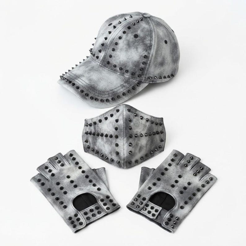 Puede incluir: Una gorra de b&eacute;isbol, una mascarilla y un par de guantes sin dedos con un acabado gris y negro lavado al &aacute;cido. Cada art&iacute;culo presenta un aspecto moteado y est&aacute; decorado con numerosas peque&ntilde;as p&uacute;as met&aacute;licas negras dispuestas en filas.