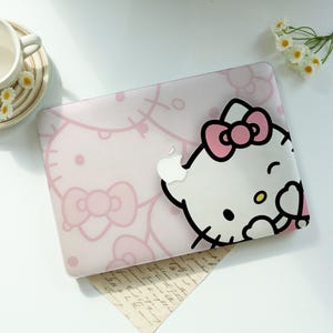 May include: A protective laptop case featuring a light pink background with a repeating Hello Kitty pattern and a large, detailed Hello Kitty character graphic on the right side, complete with a pink bow and a cutout for the Apple logo.