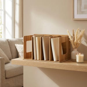 Peut inclure: Un organiseur de bureau en bois avec plusieurs compartiments, contenant plusieurs livres aux couvertures crème. L'organisateur a une découpe rectangulaire de chaque côté pour une manipulation facile. Une bougie allumée et un vase avec des fleurs séchées sont sur l'étagère.