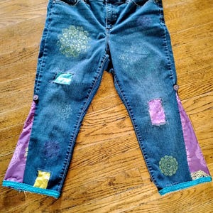 May include: Blue denim jeans with decorative patches and floral designs. The jeans feature patches of purple, yellow, and teal fabric. The sides of the jeans have purple fabric with a floral pattern and teal trim. The jeans have a button closure.