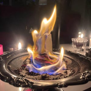 Puede incluir: Un plato plateado y ornamentado contiene incienso en llamas, con llamas amarillas, naranjas y azules. Las llamas se elevan desde un objeto blanco, rodeado de cenizas. La escena est&aacute; tenuemente iluminada, con otras velas al fondo.