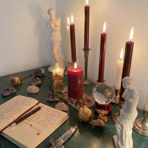 Puede incluir: Una colecci&oacute;n de objetos que incluye varias velas encendidas, tanto c&oacute;nicas como de pilar, en rojo oscuro y blanco, dos estatuas cl&aacute;sicas blancas, una bola de cristal, una daga de metal, un cuaderno con texto escrito a mano, amatistas y rosas secas.