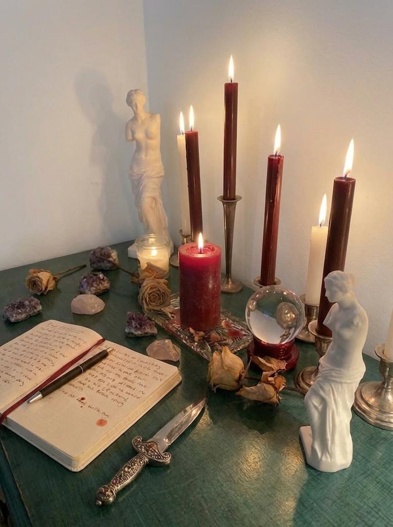 Puede incluir: Una colecci&oacute;n de objetos que incluye varias velas encendidas, tanto c&oacute;nicas como de pilar, en rojo oscuro y blanco, dos estatuas cl&aacute;sicas blancas, una bola de cristal, una daga de metal, un cuaderno con texto escrito a mano, amatistas y rosas secas.