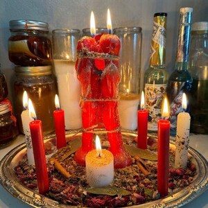 K&ouml;nnte beinhalten: Eine brennende rote Kerze in Form von zwei sich umarmenden Figuren, mit Bindfaden zusammengebunden, umgeben von anderen brennenden Kerzen in verschiedenen Gr&ouml;&szlig;en und Farben. Die Kerzen stehen auf einem silbernen Tablett mit getrockneten Kr&auml;utern und Gew&uuml;rzen, mit Gl&auml;sern und Flaschen im Hintergrund.