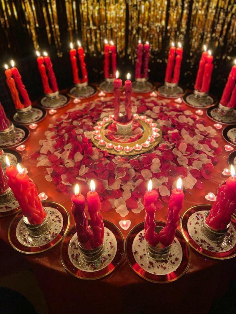 Puede incluir: Un arreglo de mesa circular con numerosos pares de velas c&oacute;nicas rojas en soportes de vidrio, rodeadas de p&eacute;talos de rosa rojos y blancos esparcidos. Peque&ntilde;as velas de t&eacute; en forma de coraz&oacute;n forman un anillo alrededor de un par central.