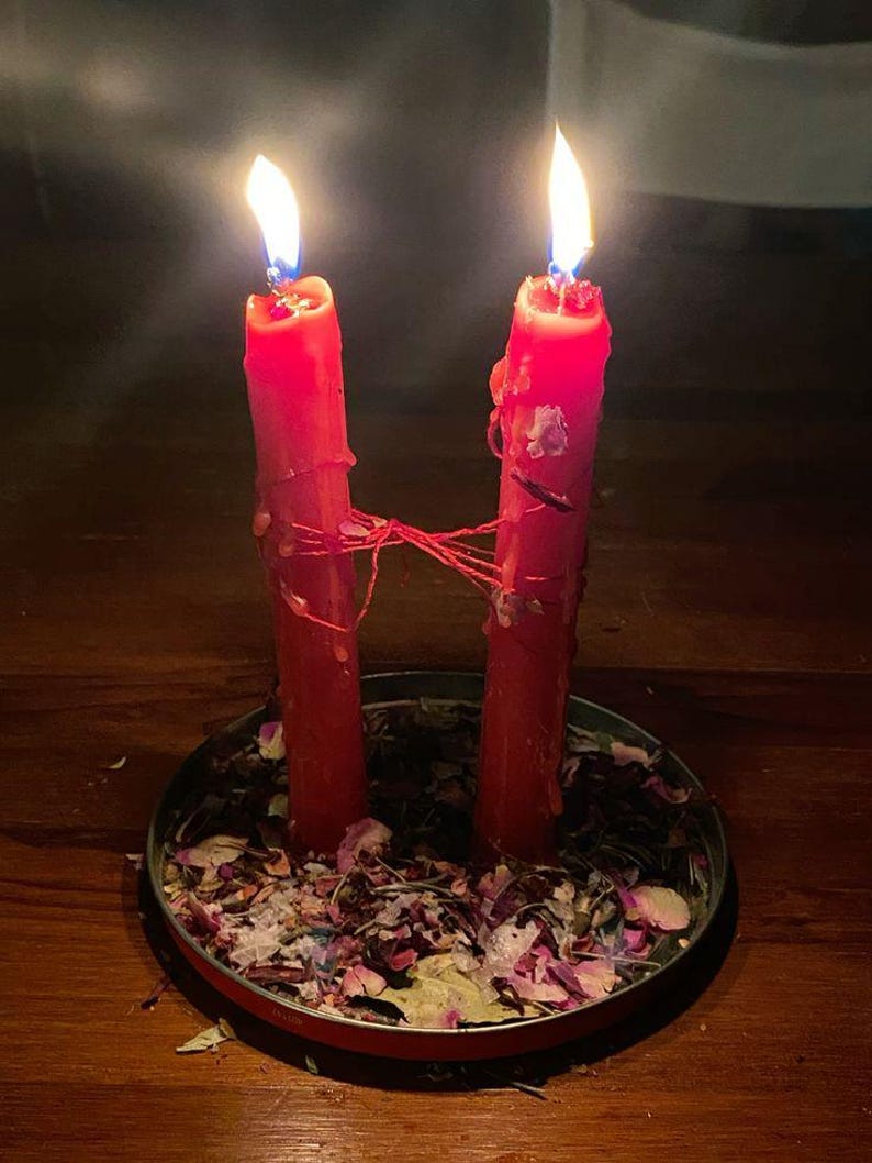 Puede incluir: Dos velas rojas encendidas, unidas por un hilo rojo, se encuentran en un plato redondo con borde rojo lleno de p&eacute;talos secos. Las velas gotean cera y las llamas brillan sobre un fondo oscuro. La imagen evoca un ritual o ceremonia.
