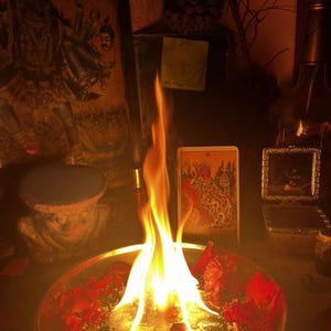 Puede incluir: Un cuenco redondo de metal lleno de agua y p&eacute;talos de rosa rojos esparcidos, con una llama brillante que se eleva desde el centro. Detr&aacute;s del cuenco hay una carta del tarot que representa una figura sentada, junto a una imagen enmarcada.