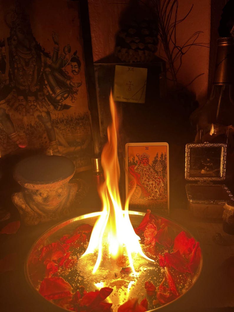 Puede incluir: Un cuenco redondo de metal lleno de agua y p&eacute;talos de rosa rojos esparcidos, con una llama brillante que se eleva desde el centro. Detr&aacute;s del cuenco hay una carta del tarot que representa una figura sentada, junto a una imagen enmarcada.