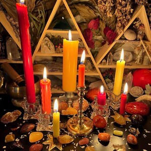 Puede incluir: Una colecci&oacute;n de velas encendidas en varios tonos de rojo, naranja y amarillo, colocadas en soportes de vidrio y metal. Las velas est&aacute;n rodeadas por numerosas piedras pulidas y cristales sobre una superficie oscura, con un estante triangular.