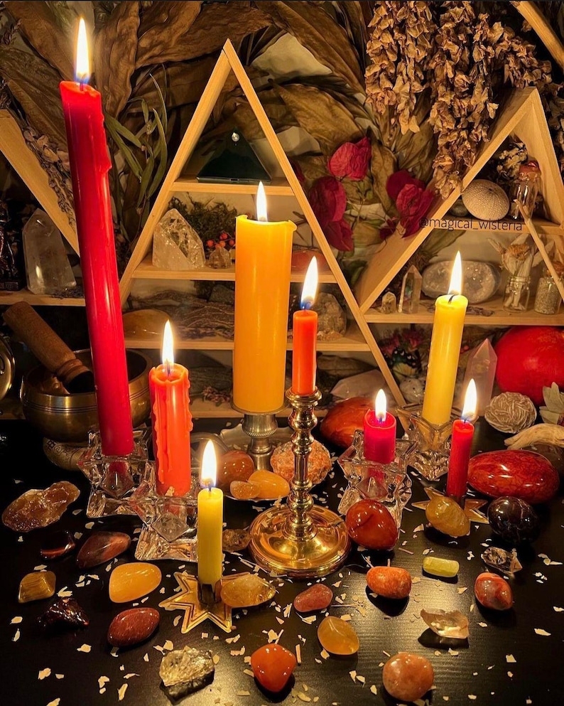 Puede incluir: Una colecci&oacute;n de velas encendidas en varios tonos de rojo, naranja y amarillo, colocadas en soportes de vidrio y metal. Las velas est&aacute;n rodeadas por numerosas piedras pulidas y cristales sobre una superficie oscura, con un estante triangular.