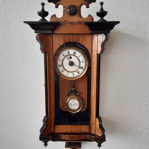 Peut inclure: Une horloge murale ancienne en bois avec un pendule. L'horloge a un cadre en bois brun clair avec des accents noirs et un cadran blanc avec des chiffres romains noirs. L'horloge a un dessus et un bas décoratifs.