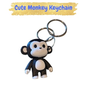 Puede incluir: Un llavero de mono en blanco y negro con anillos plateados. El llavero tiene el texto "Cute Monkey Keychain" en letras azules sobre un fondo dorado.