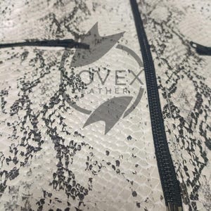 K&ouml;nnte beinhalten: Nahaufnahme einer Jacke mit Schlangenmuster in Wei&szlig;-, Grau- und Schwarzt&ouml;nen. Die Jacke hat einen schwarzen Rei&szlig;verschluss und den Text "OVEX LEATHER". Die Textur des Materials ist sichtbar.