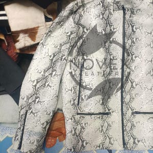K&ouml;nnte beinhalten: Wei&szlig;-graue Jacke mit Schlangenmuster, schwarzem Rei&szlig;verschluss und Taschen. Die Jacke hat einen Stehkragen und eine taillierte Passform. Der Markenname "NOVE LEATHER" ist sichtbar.