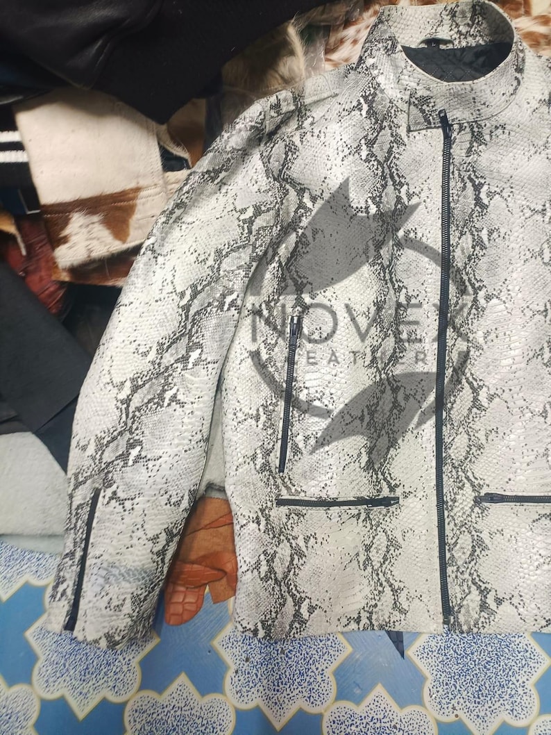 K&ouml;nnte beinhalten: Wei&szlig;-graue Jacke mit Schlangenmuster, schwarzem Rei&szlig;verschluss und Taschen. Die Jacke hat einen Stehkragen und eine taillierte Passform. Der Markenname "NOVE LEATHER" ist sichtbar.