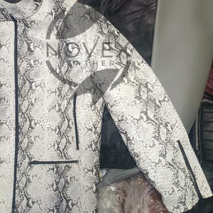 K&ouml;nnte beinhalten: Wei&szlig;-graue Jacke mit Schlangenmuster, Rei&szlig;verschluss und schwarzen Zierleisten. Die Jacke hat einen Stehkragen und zwei Vordertaschen. Der Markenname "NOVEATHER" ist sichtbar. Andere Lederjacken sind im Hintergrund.