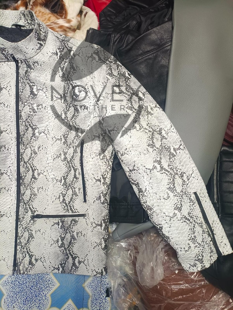 K&ouml;nnte beinhalten: Wei&szlig;-graue Jacke mit Schlangenmuster, Rei&szlig;verschluss und schwarzen Zierleisten. Die Jacke hat einen Stehkragen und zwei Vordertaschen. Der Markenname "NOVEATHER" ist sichtbar. Andere Lederjacken sind im Hintergrund.