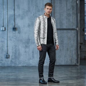 K&ouml;nnte beinhalten: Eine wei&szlig;-graue Jacke mit Schlangenmuster und schwarzem Besatz. Die Jacke wird &uuml;ber einem schwarzen T-Shirt getragen und mit dunkelgrauen Jeans und schwarzen Stiefeln kombiniert. Das Outfit wird durch einen schwarzen G&uuml;rtel erg&auml;nzt.