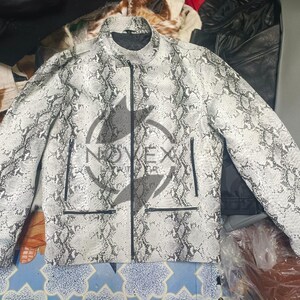K&ouml;nnte beinhalten: Eine wei&szlig;-graue Jacke mit Schlangenmuster, schwarzem Rei&szlig;verschluss und Taschen. Die Jacke hat einen Stehkragen und Rei&szlig;verschl&uuml;sse an den &Auml;rmeln. Die Jacke ist mittelschwer und scheint aus Leder zu sein.