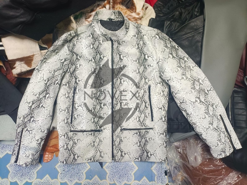 K&ouml;nnte beinhalten: Eine wei&szlig;-graue Jacke mit Schlangenmuster, schwarzem Rei&szlig;verschluss und Taschen. Die Jacke hat einen Stehkragen und Rei&szlig;verschl&uuml;sse an den &Auml;rmeln. Die Jacke ist mittelschwer und scheint aus Leder zu sein.