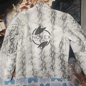 K&ouml;nnte beinhalten: Eine wei&szlig;e und graue Lederjacke mit Schlangenmuster und langen &Auml;rmeln. Auf der R&uuml;ckseite der Jacke befindet sich ein schwarzes Logo mit der Aufschrift "NOVEX LEATHER" in einem kreisf&ouml;rmigen Design. Die Jacke ist neutralfarben und hat ein klassisches Design.