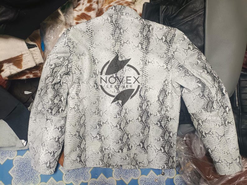 K&ouml;nnte beinhalten: Eine wei&szlig;e und graue Lederjacke mit Schlangenmuster und langen &Auml;rmeln. Auf der R&uuml;ckseite der Jacke befindet sich ein schwarzes Logo mit der Aufschrift "NOVEX LEATHER" in einem kreisf&ouml;rmigen Design. Die Jacke ist neutralfarben und hat ein klassisches Design.