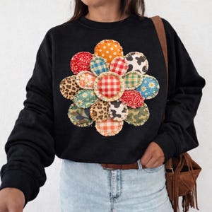 May include: Black sweatshirt featuring a colorful, patchwork flower design. The flower is composed of various patterned fabric circles, including polka dots, gingham, and animal prints. The sweatshirt has a crew neck and long sleeves.